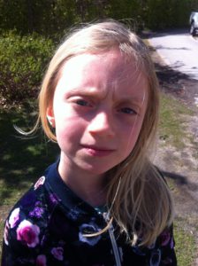 Elsa står utomhus i solen. Hon har långt blont hår och rynkar pannan.