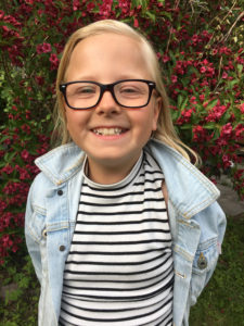 Felicia ler stort i sina glasögon framför rosa blommor.