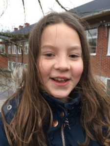 Lea ståt utomhus med en röd byggnad i bakgrunden. Hon har svart jacka och brunt långt hår som blåser i vinden.