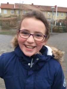 Elina ler stort mot kameran. Hon har glasögon och brunt hår i hästsvans.