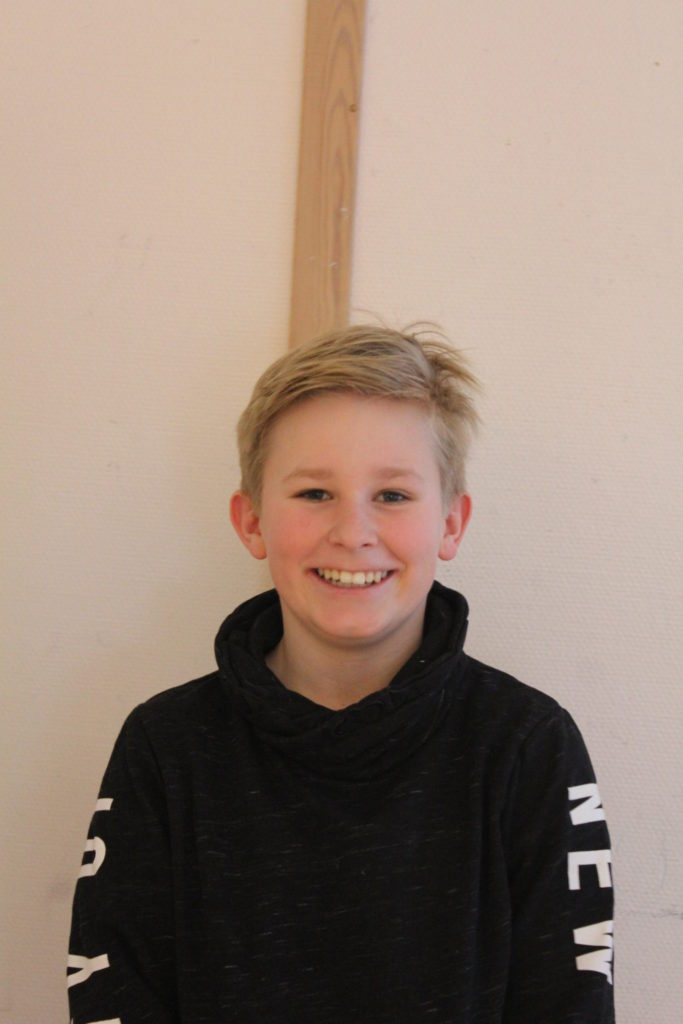 Gustav står framför en vit vägg. Han har svart tröja och blont rufsigt hår. Han ler stort.