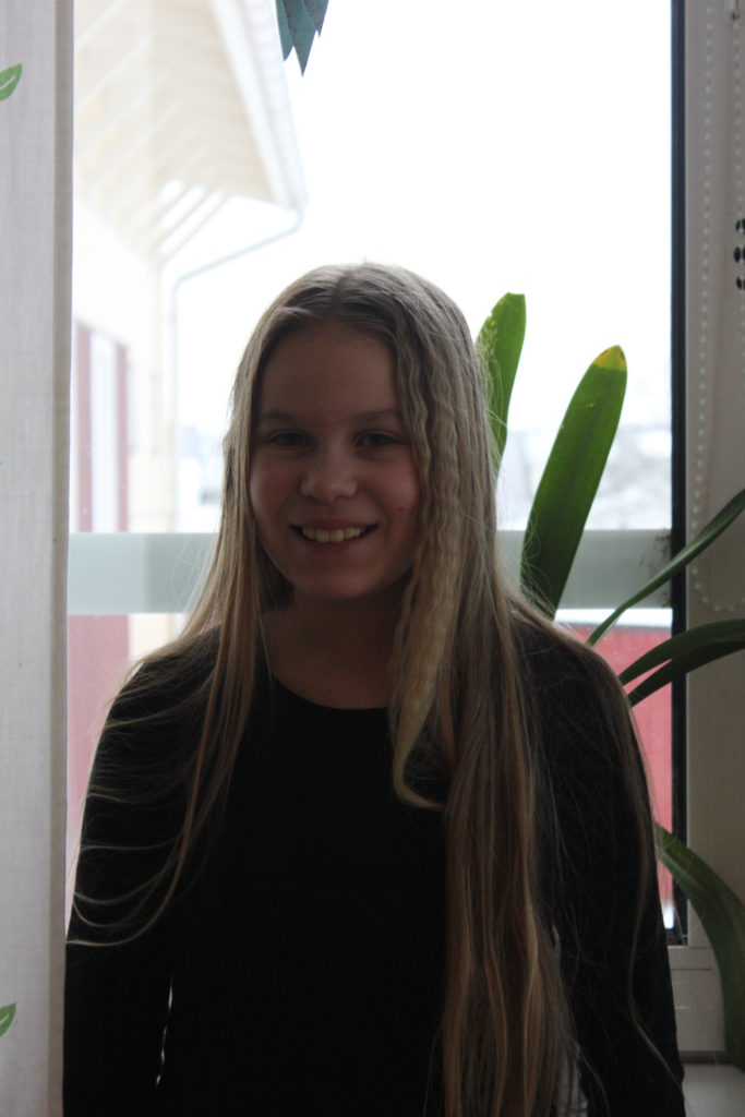 Molly står framför ett fönster. Hon har en mörk tröja och långt blont hår. Hon tittar in i kameran och ler.