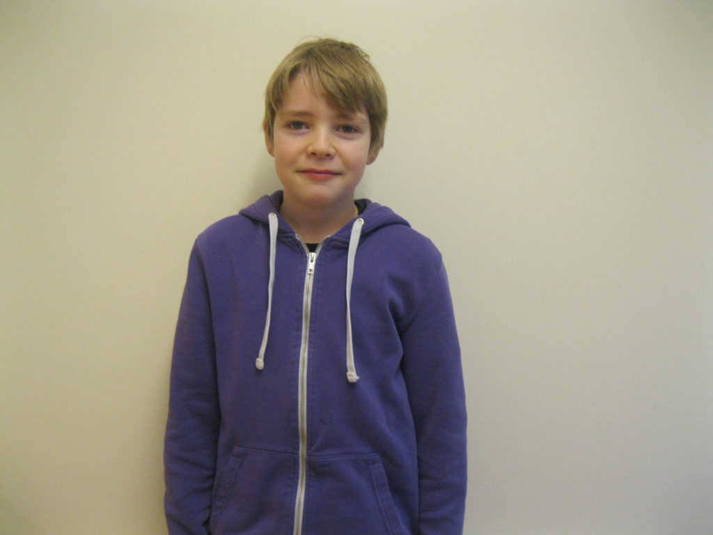 Titus står framför en vit vägg. Han har en blå munktröja med vita snören. Han har kort blont hår.