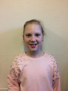 Alva står framför en vit vägg. Hon har en rosa tröja med pärlor på och blont hår i tofs. Hon ler mot kameran.