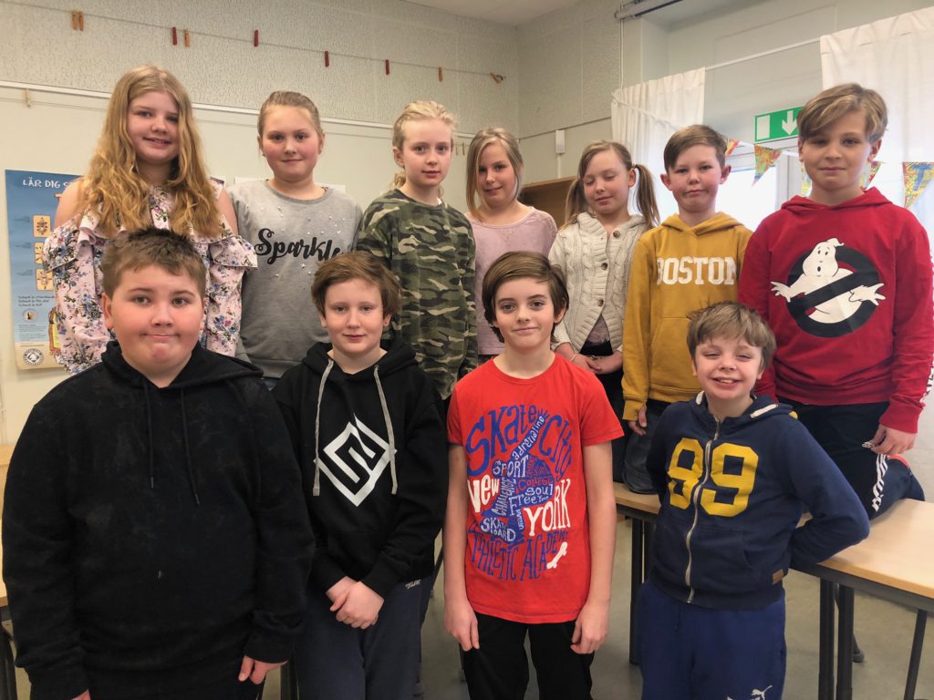 Klass 4D står i sitt klassrum. De är uppställda i två rader och tittar ni i kameran. De är 12 personer som alla har olika kläder.