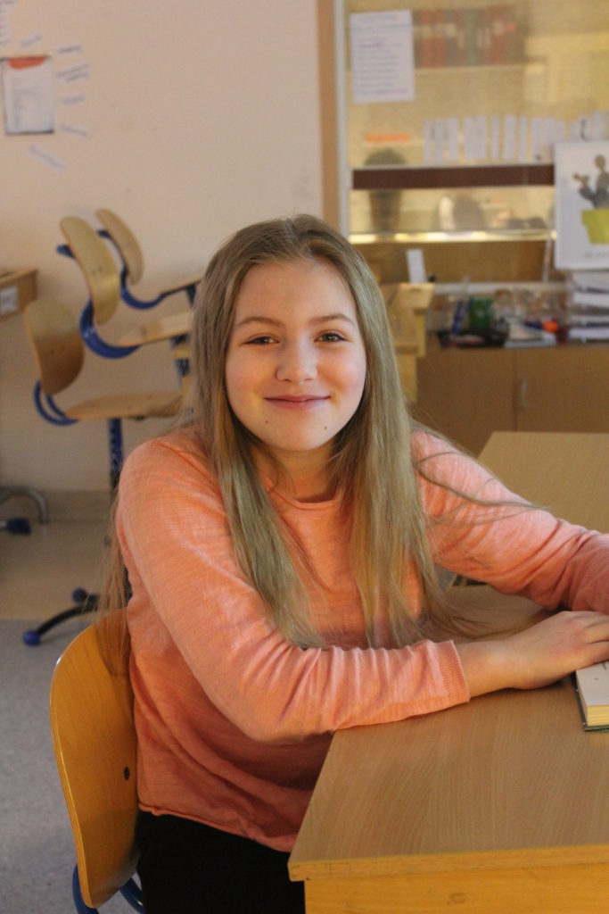 Agnes sitter vid en skolbänk och vrider sig åt sidan för att titta in i kameran. Hon har långt blont hår och en aprikosfärgad tröja.