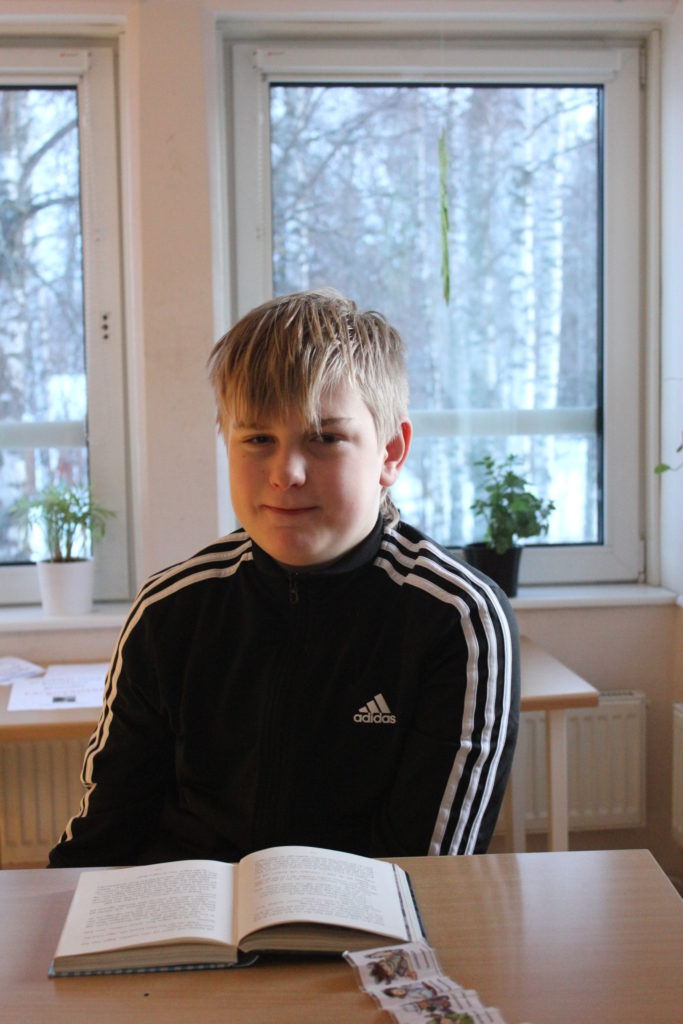 Oscar sitter bakom en skolbänk med ett fönster bakom sig. Han har kort blont hår med lugg som ligger ner i pannan, och en svarta adidaströja med vita ränder på armarna.