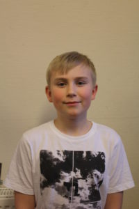 Erik står framför en vit vägg. Han har en vit t-shirt med svart tryck på. Han har blont hår med lugg som ligger ner över pannan.