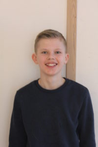 Adam står framför en vit vägg. Han har en blå längärmad tröja och blont hår med lugg som ligger åt sidan. Han ler mot kameran.