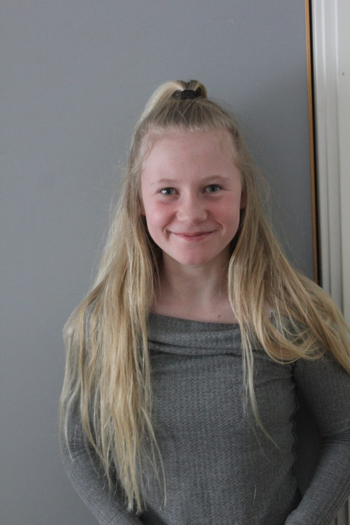 Emilia står framför en grå vägg. Hon har en grå tröja. Hon har långt blont hår där det främst håret är uppsatt i en tofs på huvudet.
