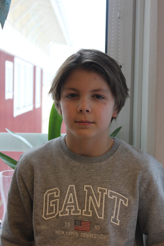 Adam står framför ett fönster. Han har en grå tröja och halvlångt hår med en mittbena.