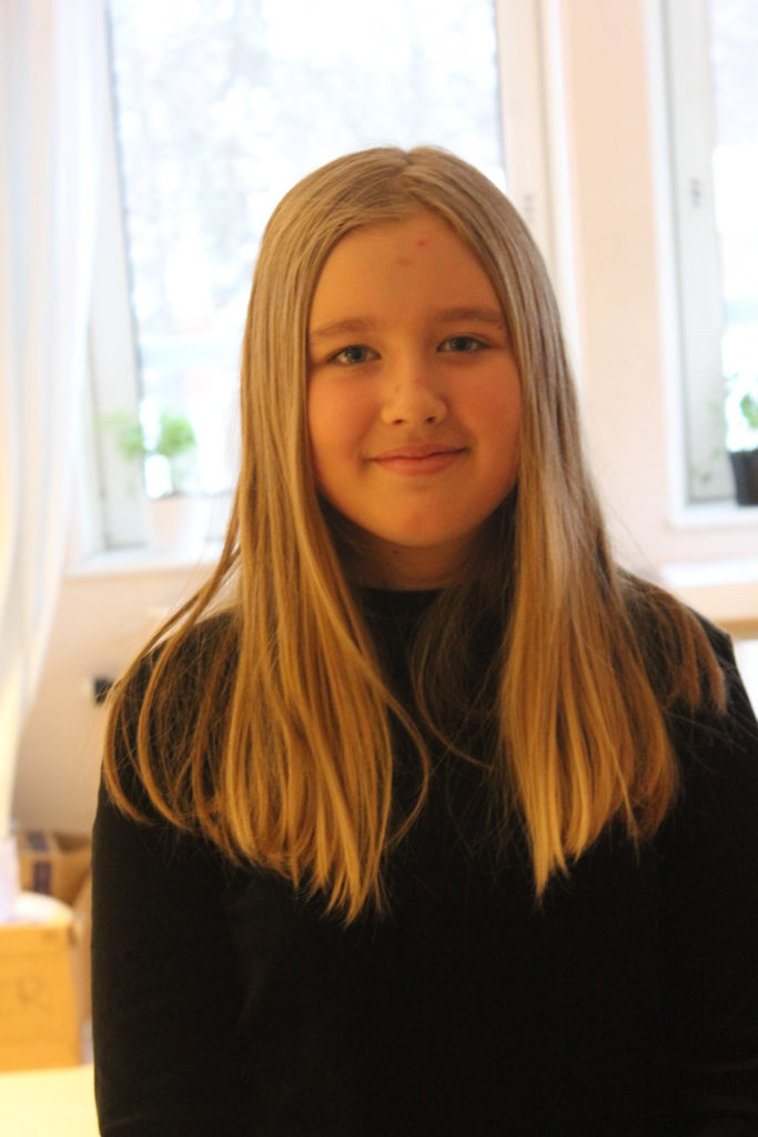 Amanda står i ett ljust rum. Hon tittar in i kameran. Hon haren svart tröja och långt ljust hår som hänger ner över axlarna.