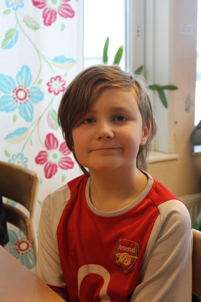 Emil sitter vid en skolbänk. Han har halvlångt brunt hår och en röd fotbollströja.