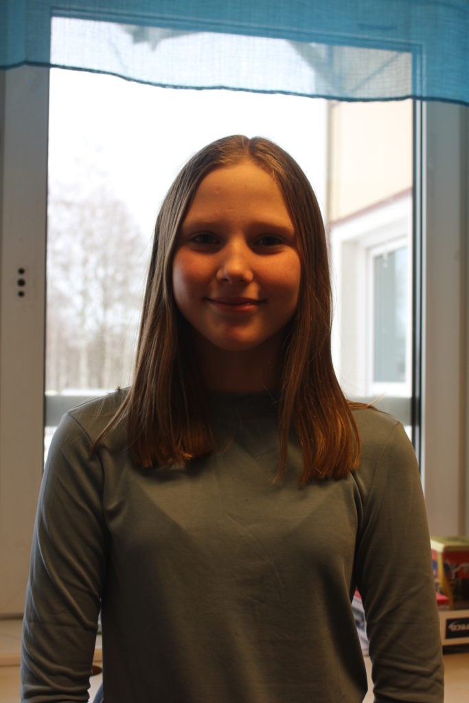 Tyra står framför ett fönster. Hon har en långärmad tröja på sig och axellångt hår i en mittbena.