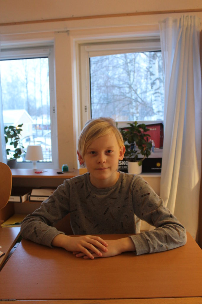 Olivers sitter bakom en skolbänk. Han har långärmad tröja och blont hår med lugg som ligger åt sidan. Han tittar bestämt in i kameran.