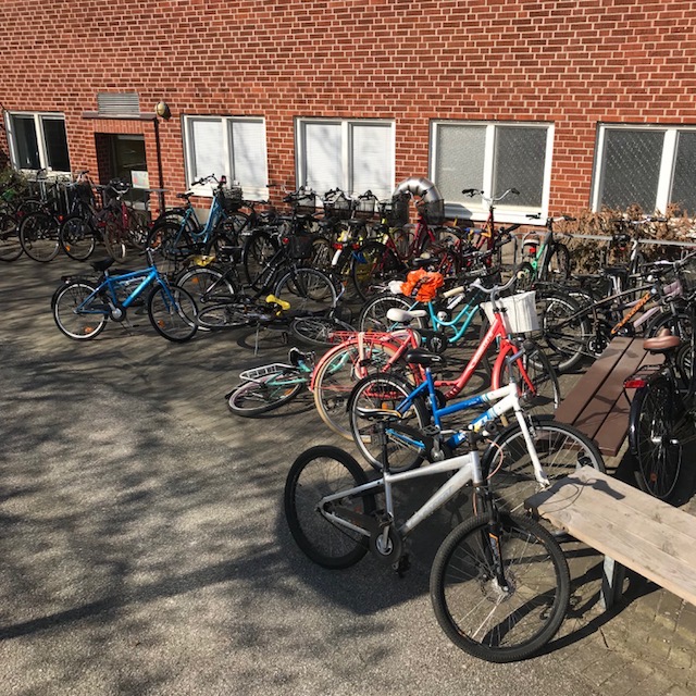 En cykelparkering intill en vägg som är helt full av cyklar som ligger välta på marken och många som står utanför cykelställen.