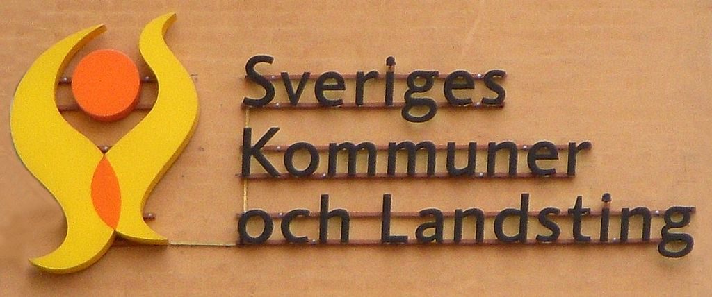 En skylt med en gul och orange symbol som ser lite ut som en eldsflamma och lite som en gubbe. Bredvid står det "Sveriges kommuner och landsting".