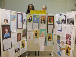 Två skolelever visar upp en vägg med uppsatta skolarbeten över historiska kvinnor under rubriken: Rebellkvinnor.