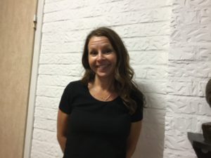 En person med långt brunt hår ler mot kameran framför en vit vägg.