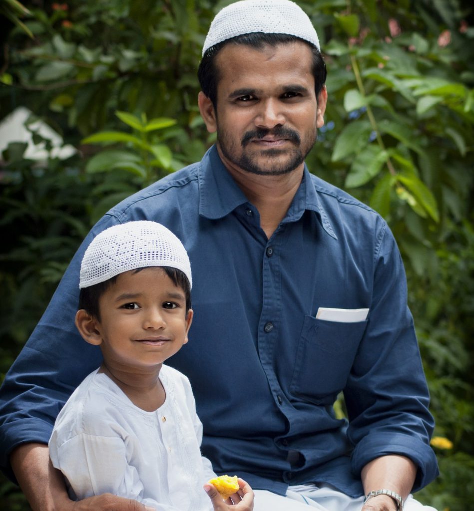 En pappa sitter och håller om sitt barn. Båda har vita små virkade mössor på sig. Barnet äter en bot frukt och ser glad ut.