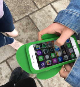 En hand håller i en smartphone med grönt mobilskal i plast. I bakgrunden syns markens stenplattor och två personers skor.