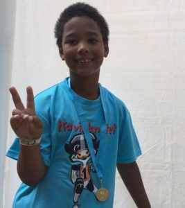 Tijan håller upp fingrarna i ett V-märke. Om halsen hänger hans medalj.