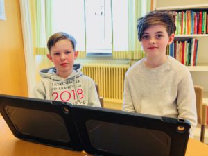 De två skolelever som skrivit reportaget sitter vid skrivbordet i klassrummet med två skärmar framför sig.