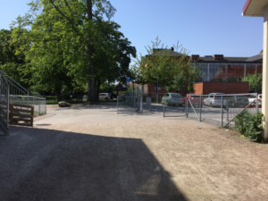 Bild på skolgårdens öppna ingång med ett träd och bilparkering och grusgång.