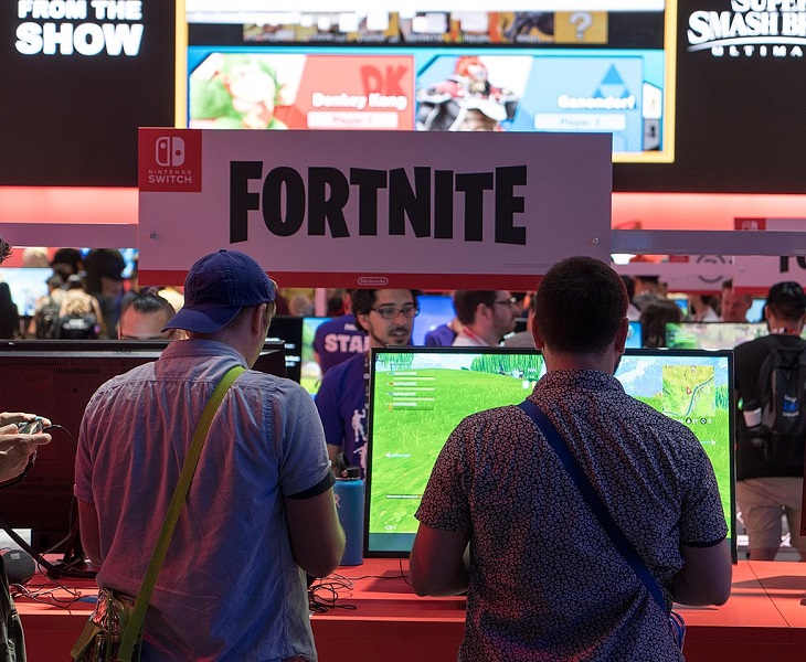 Två spelare står framför en dataskärm och spelar. Bortanför dem syns en skylt där det står Fortnite.