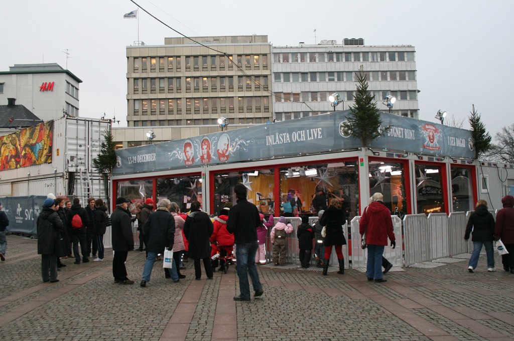 En liten byggnad står på ett torg. Den har glasväggar och längst upp sitter reklam för Musikhjälpen. Utanför buren står det massa människor.