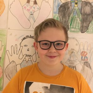 Elliot står framför en vägg med teckningar. Han har kort hår och glasögon. Han ser glad ut.