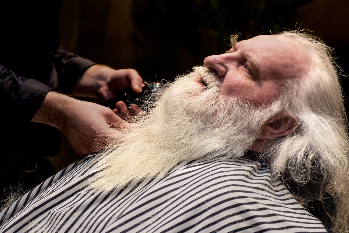 En man ligger ner i en frisörstol. Han har ett stort skägg och mustasch. En person håller fram en rakapparat mot skägget.