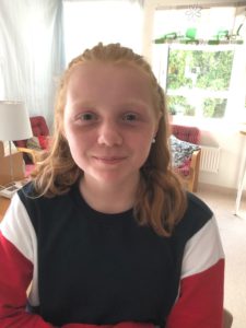 Julia kollar in i kameran och ler. Solen lyser in genom fönstret i bakgrunden och man kan se löven på trädet.