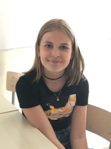 Wilma sitter i sin skolbänk. Hon har armarna i knät och ler när hon tittar in i kameran.